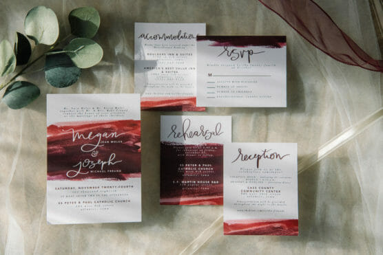A set of wedding stationery on a table, including invitation, RSVP, accommodation, rehearsal, and reception cards, featuring elegant calligraphy and red watercolor accents, with eucalyptus leaves and a sheer burgundy fabric for decoration. by Justin Salem Meyer