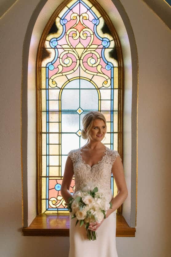 A bride in a lace wedding gown holds a white bouquet and stands in front of a colorful stained glass window, softly lit by natural light. by Justin Salem Meyer