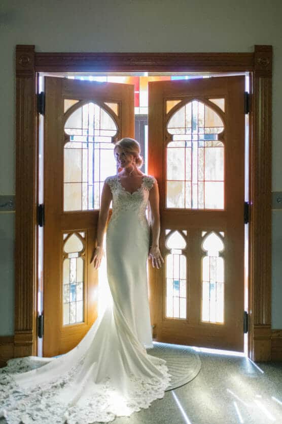 A bride in a fitted, lace wedding gown stands in front of large wooden doors with stained glass panels, illuminated by sunlight streaming in from behind. by Justin Salem Meyer