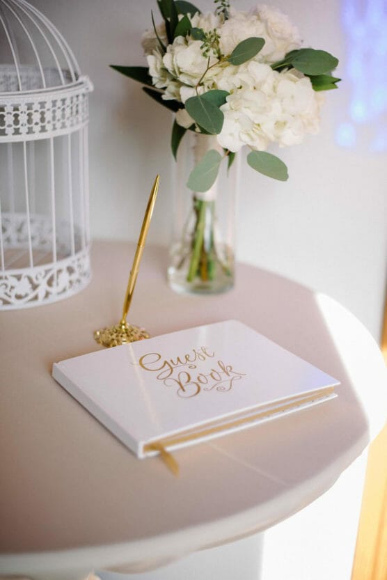 A white guest book with gold lettering and a gold pen sits on a round table next to a glass vase of white flowers and a white decorative birdcage. by Justin Salem Meyer