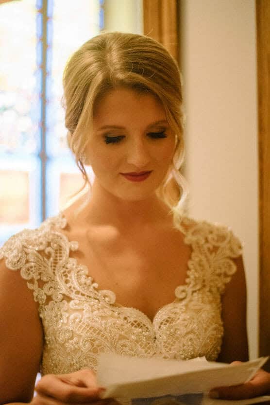 A bride in an elegant lace wedding dress reads a letter, standing indoors with soft natural light and a blurred stained glass window in the background. by Justin Salem Meyer