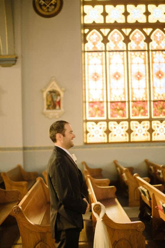 A man in a suit stands smiling in a church pew, facing right. Sunlight filters through a large stained glass window behind him, casting a warm glow inside the church. by Justin Salem Meyer