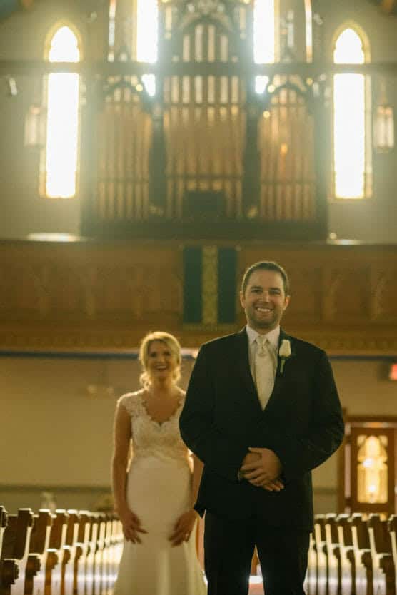 A groom in a dark suit stands smiling in a sunlit church aisle, while a bride in a white gown stands behind him, also smiling, with glowing stained glass windows in the background. by Justin Salem Meyer