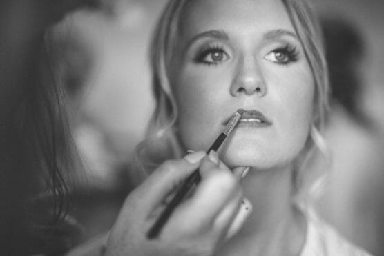 A close-up, black-and-white photo of a woman having lipstick applied to her lips by another person, focusing on her face and the makeup brush. by Justin Salem Meyer