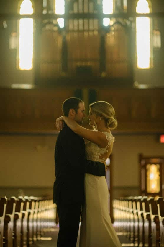 A bride and groom stand close together, embracing and gazing at each other in the aisle of a dimly lit church, with sunlight streaming through tall stained glass windows behind them. by Justin Salem Meyer