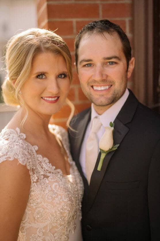A bride and groom stand close together, smiling at the camera. The bride wears an embellished lace wedding dress, and the groom wears a black suit with a white tie and rose boutonniere. They are outdoors near a brick wall. by Justin Salem Meyer