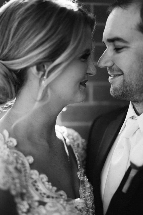 A bride and groom stand close together, smiling at each other on their wedding day. The image is in black and white, highlighting their happy expressions and elegant attire. by Justin Salem Meyer