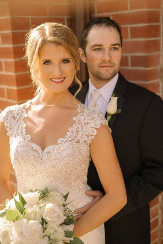 A bride in a lace wedding dress holding a bouquet of white flowers stands in front of a groom in a dark suit with a white rose boutonniere. They pose together, smiling, in front of a brick wall. by Justin Salem Meyer