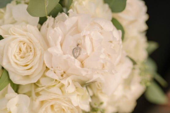 A close-up of a bouquet of white flowers, including roses and peonies, with a small silver heart-shaped pendant nestled among the petals. by Justin Salem Meyer
