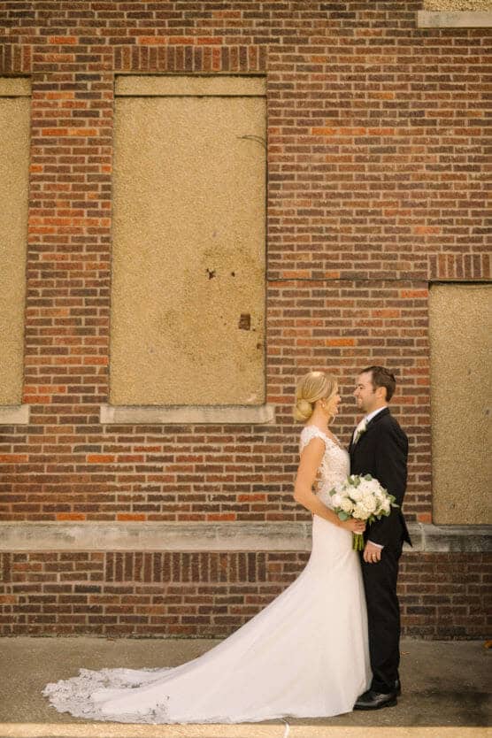 A bride in a white gown holding a bouquet stands facing a groom in a black suit. They smile at each other in front of a brick wall with boarded windows. by Justin Salem Meyer