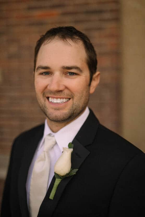 A man in a black suit and cream tie smiles at the camera, wearing a white rose boutonniere. He stands in front of a blurred brick wall background. by Justin Salem Meyer