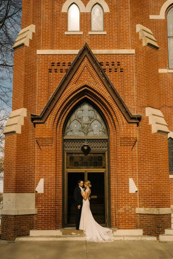 A bride and groom stand embracing in front of the large arched entrance of a red brick church, with sunlight illuminating the scene. by Justin Salem Meyer