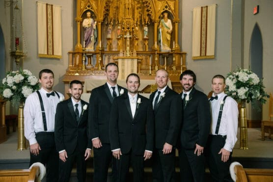 Seven men dressed in formal attire pose and smile together inside a church with an ornate golden altar and floral arrangements in the background. by Justin Salem Meyer