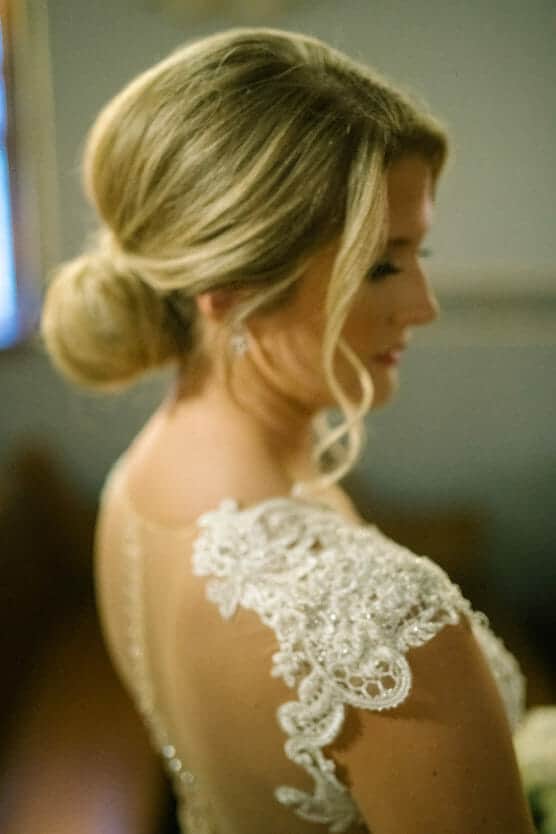 A woman in a lace wedding dress with an intricate updo hairstyle is shown from the side, her face softly out of focus and turned slightly away from the camera. by Justin Salem Meyer