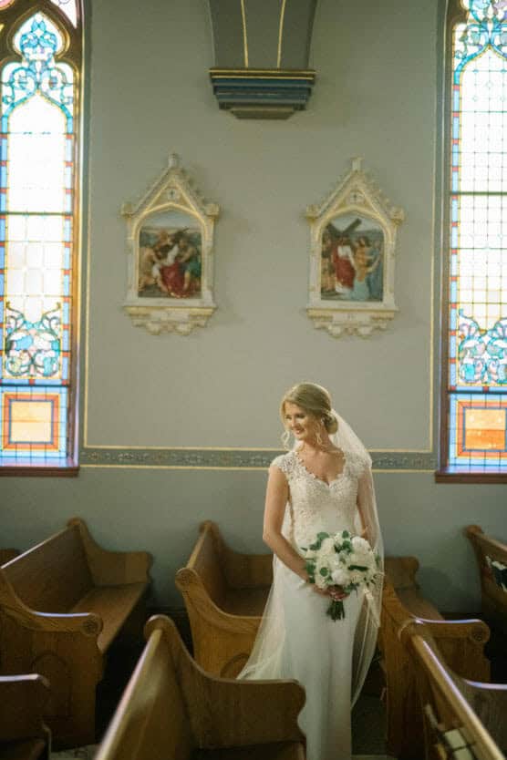 A bride in a white dress and veil holds a bouquet of flowers while standing in a church pew, with stained glass windows and religious artwork in the background. by Justin Salem Meyer