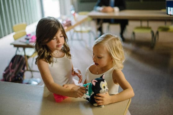 Two young girls in white tops play with a plush cat toy at a table in a classroom. One girl brushes the toy while the other watches. School desks and chairs are visible in the background. by Justin Salem Meyer