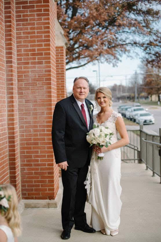 A bride in a white dress holding a bouquet stands smiling beside a man in a black suit and tie outside a brick building. Trees with autumn leaves and parked cars line the street in the background. by Justin Salem Meyer