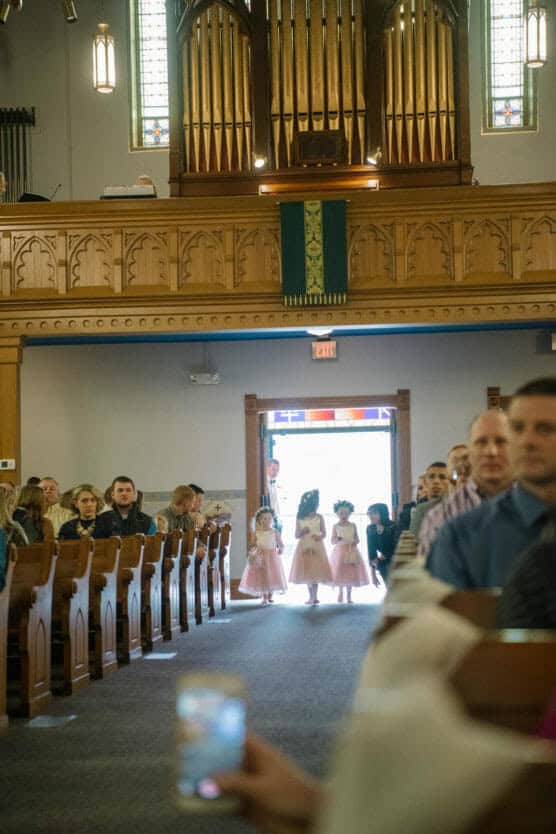 Four young girls in pink dresses and flower crowns walk down the aisle of a church filled with seated guests. Stained glass windows and a large organ are visible in the background. Light streams in from the open doorway behind them. by Justin Salem Meyer