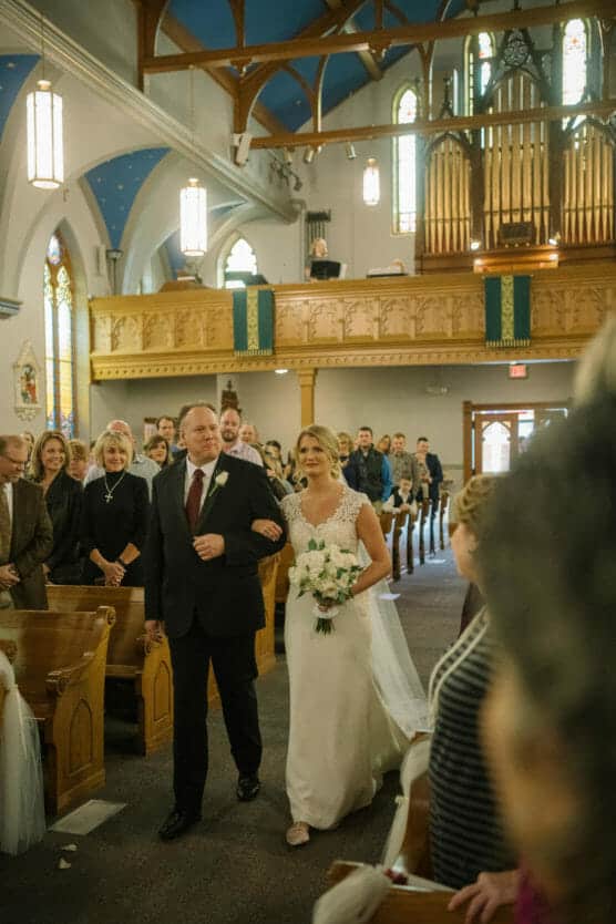 A bride in a white gown holding a bouquet walks down the aisle arm-in-arm with a man in a suit inside a church, while guests watch from the pews. The church has stained glass windows and a large organ. by Justin Salem Meyer