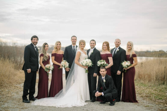 A wedding party poses outdoors on a gravel path near tall grass. The bride in a white gown and the groom in a black suit stand at the center, surrounded by bridesmaids in burgundy dresses and groomsmen in black suits, all holding bouquets. by Justin Salem Meyer