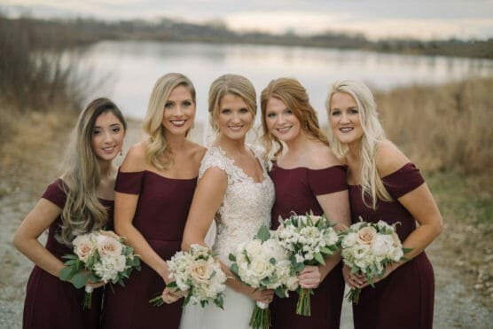 Five women in formal dresses pose outdoors by a lake. The woman in the center wears a white wedding gown, while the others wear matching burgundy dresses and hold bouquets of white and blush flowers. The background is natural and serene. by Justin Salem Meyer