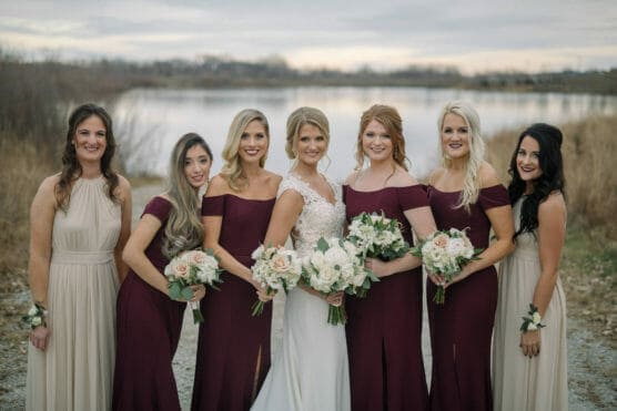 A bride in a white gown stands with six bridesmaids in burgundy and beige dresses, all holding bouquets, posing outdoors by a lake with overcast skies and dry grass in the background. by Justin Salem Meyer