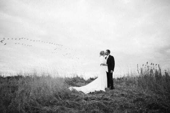 A bride and groom stand in a grassy field, sharing a kiss under a cloudy sky, with a flock of birds flying in the background. The photo is black and white, creating a timeless, romantic atmosphere. by Justin Salem Meyer