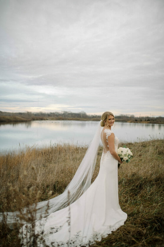 A bride in a white gown with a long veil stands on grass near a calm lake under a cloudy sky, holding a bouquet of white flowers and looking over her shoulder at the camera. by Justin Salem Meyer