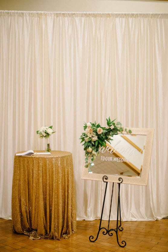 A gold sequin-covered table with a small bouquet and guest book stands beside a mirror sign reading Welcome to our wedding, decorated with greenery and white flowers, set against a white draped backdrop. by Justin Salem Meyer