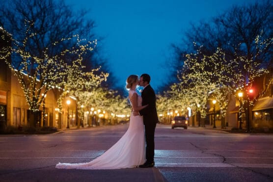 A bride and groom stand close together, facing each other in the middle of an empty street at dusk, surrounded by trees adorned with glowing string lights. by Justin Salem Meyer