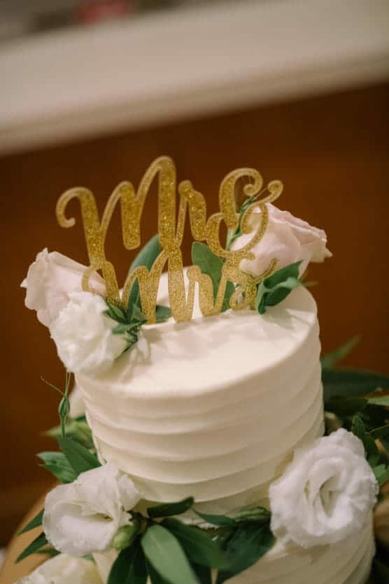 A white tiered cake decorated with white flowers and green leaves, topped with a gold Mr & Mrs sign. by Justin Salem Meyer