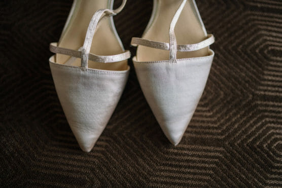 A close-up of a pair of elegant, light-colored pointed-toe shoes with thin straps, placed side by side on a patterned dark brown fabric surface. by Justin Salem Meyer