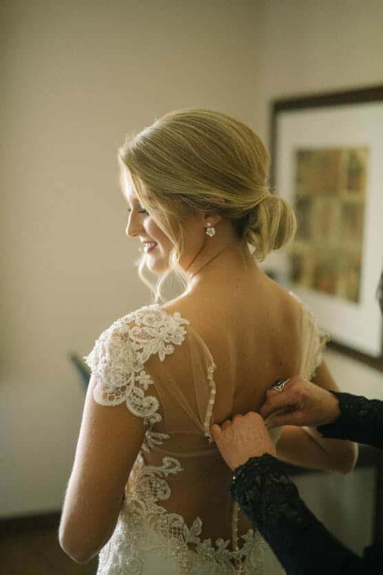 A bride with blonde hair styled in a low bun smiles as someone helps button the back of her lace wedding dress in a softly lit room. by Justin Salem Meyer