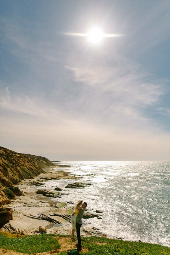 A couple stands on a grassy cliff overlooking the ocean, embracing under a bright sun and blue sky, with rocky shoreline and waves stretching into the distance. by Justin Salem Meyer