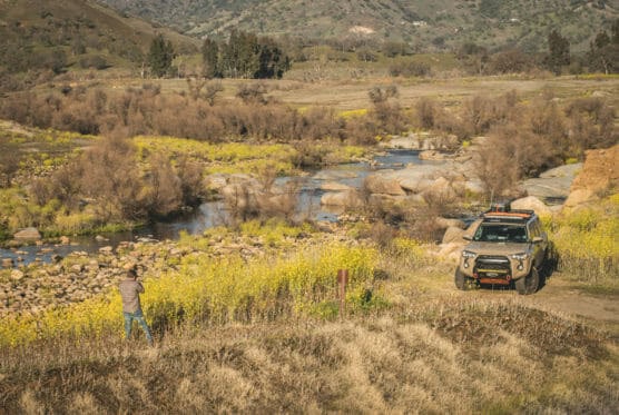 A person takes photos near a stream surrounded by yellow wildflowers, with a rugged SUV parked nearby on a dirt path; rocky hills and trees are visible in the background. by Justin Salem Meyer