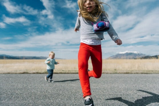 A young girl in red pants runs toward the camera on a rural road, with a younger child following behind. The sky is blue with clouds, and mountains are visible in the background. by Justin Salem Meyer