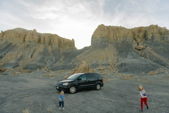 Two children play in the foreground near a black minivan parked on rocky ground, with large beige cliffs rising in the background under a partly cloudy sky. by Justin Salem Meyer