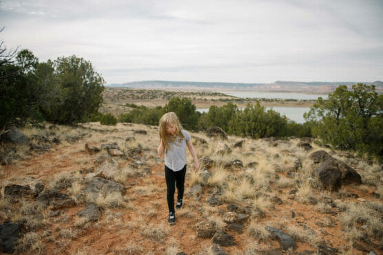 A young girl with blonde hair walks on a rocky, grassy trail in a dry landscape with scattered bushes and distant water under a cloudy sky. by Justin Salem Meyer