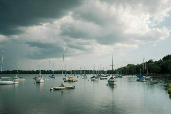 Sailboats anchored on a calm lake under a cloudy sky, with trees lining the distant shore. The water reflects the boats and muted light from the overcast atmosphere. by Justin Salem Meyer