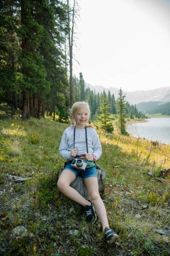 A young girl sits on a tree stump in a scenic, grassy area near a lake and pine trees, smiling and holding a camera with mountains visible in the background. by Justin Salem Meyer
