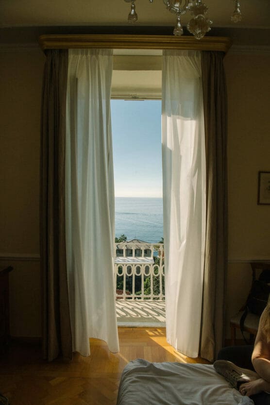 Sunlit room with open white curtains revealing a balcony overlooking the sea and a white building below; part of a bed and a person’s arm are visible in the foreground. by Justin Salem Meyer