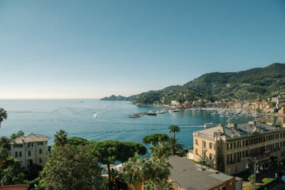A coastal town with colorful buildings, lush green hills, and a marina filled with boats beside a calm, blue sea under a clear sky. by Justin Salem Meyer