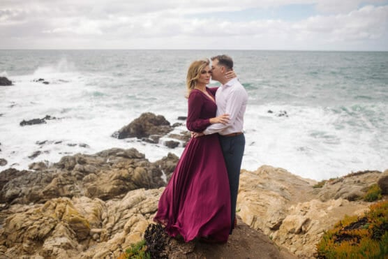 A couple stands on rocky cliffs by the ocean. The woman wears a flowing burgundy dress, while the man wears a white shirt and dark pants. Waves crash against the rocks under a cloudy sky. by Justin Salem Meyer