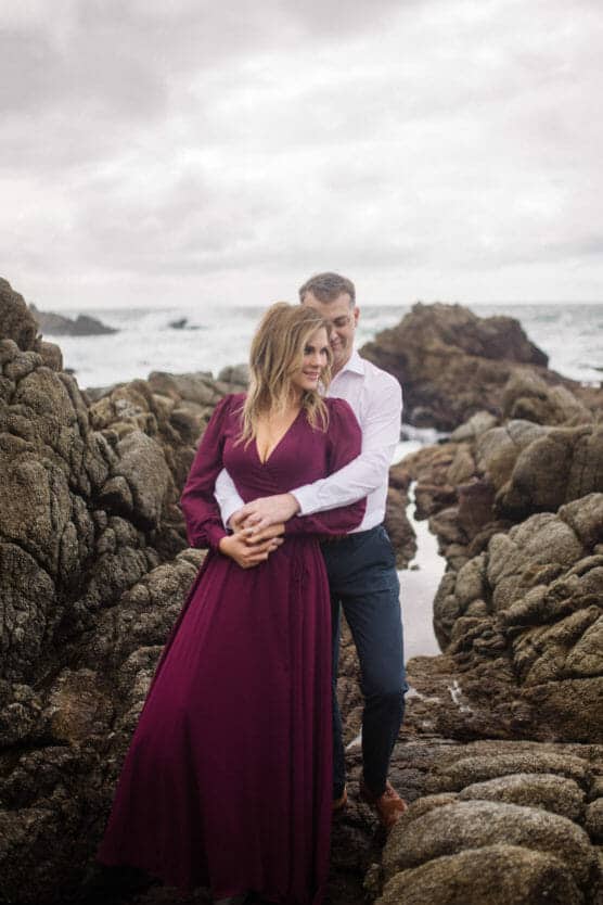 A couple stands on rocky terrain by the ocean. The woman wears a long burgundy dress, and the man stands behind her in a white shirt and dark trousers, embracing her and smiling. The sky is cloudy and the sea is visible in the background. by Justin Salem Meyer