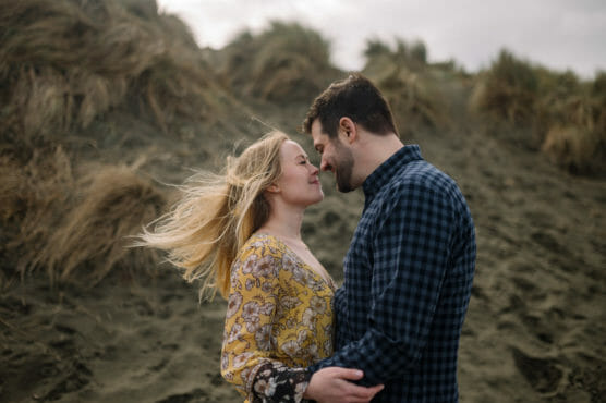 A couple stands close together on a sandy beach with grassy dunes behind them, gazing into each others eyes and smiling. The womans blonde hair blows in the wind as she wears a yellow floral dress; the man wears a blue plaid shirt. by Justin Salem Meyer