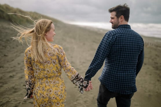 A couple holds hands and walks along a sandy beach. The woman, in a yellow floral dress, looks back smiling at the man, who wears a blue checkered shirt. The weather is cloudy and windy, with waves in the background. by Justin Salem Meyer