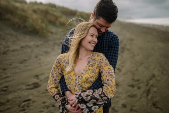 A smiling couple stands on a sandy beach. The woman, wearing a yellow floral dress, is embraced from behind by the man, who wears a blue plaid shirt. Wind blows her hair, and dunes are visible in the background. by Justin Salem Meyer