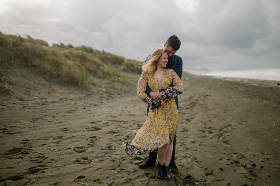 A couple stands on a sandy beach on a cloudy day. The woman, wearing a yellow floral dress, smiles as the man, in a blue shirt, hugs her from behind. Grass-covered dunes and the ocean are visible in the background. by Justin Salem Meyer