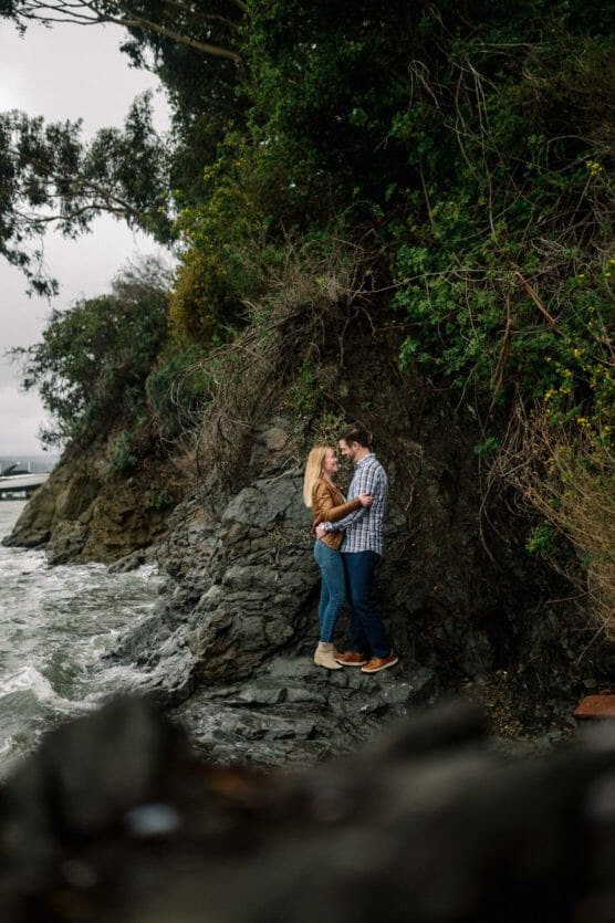 A couple stands close together on rocky terrain by the waters edge, surrounded by lush greenery and overhanging trees under a cloudy sky. by Justin Salem Meyer