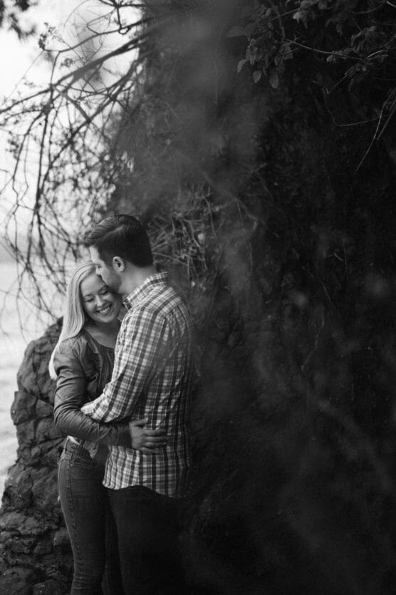 A couple stands close together outdoors near a rocky wall. The woman smiles brightly as the man embraces her, both dressed casually. The scene is in black and white, with twigs and foliage visible in the background. by Justin Salem Meyer